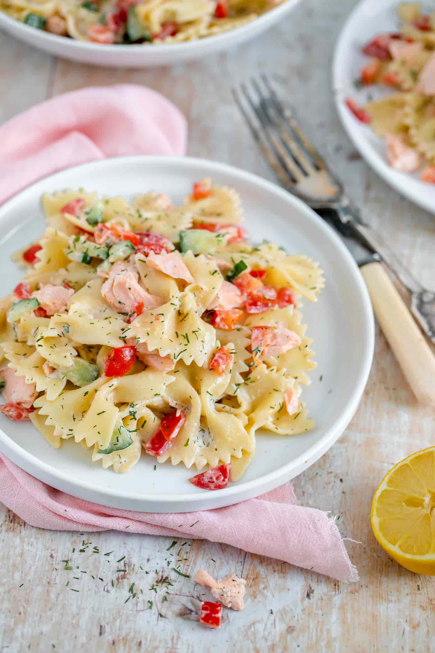 salmon pasta salad on a white plate sat on a pink napkin. A knife and fork sit to the side on an anglewith a lemon half and a corner of another 2 plates of salmon pasta salad just visible in the top right corner and the top left corner