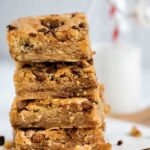 chocolate chip blondies stacked on top of each other with crumbs and chocolate chips scattered around them. A milk bottle with a red striped star is in the background