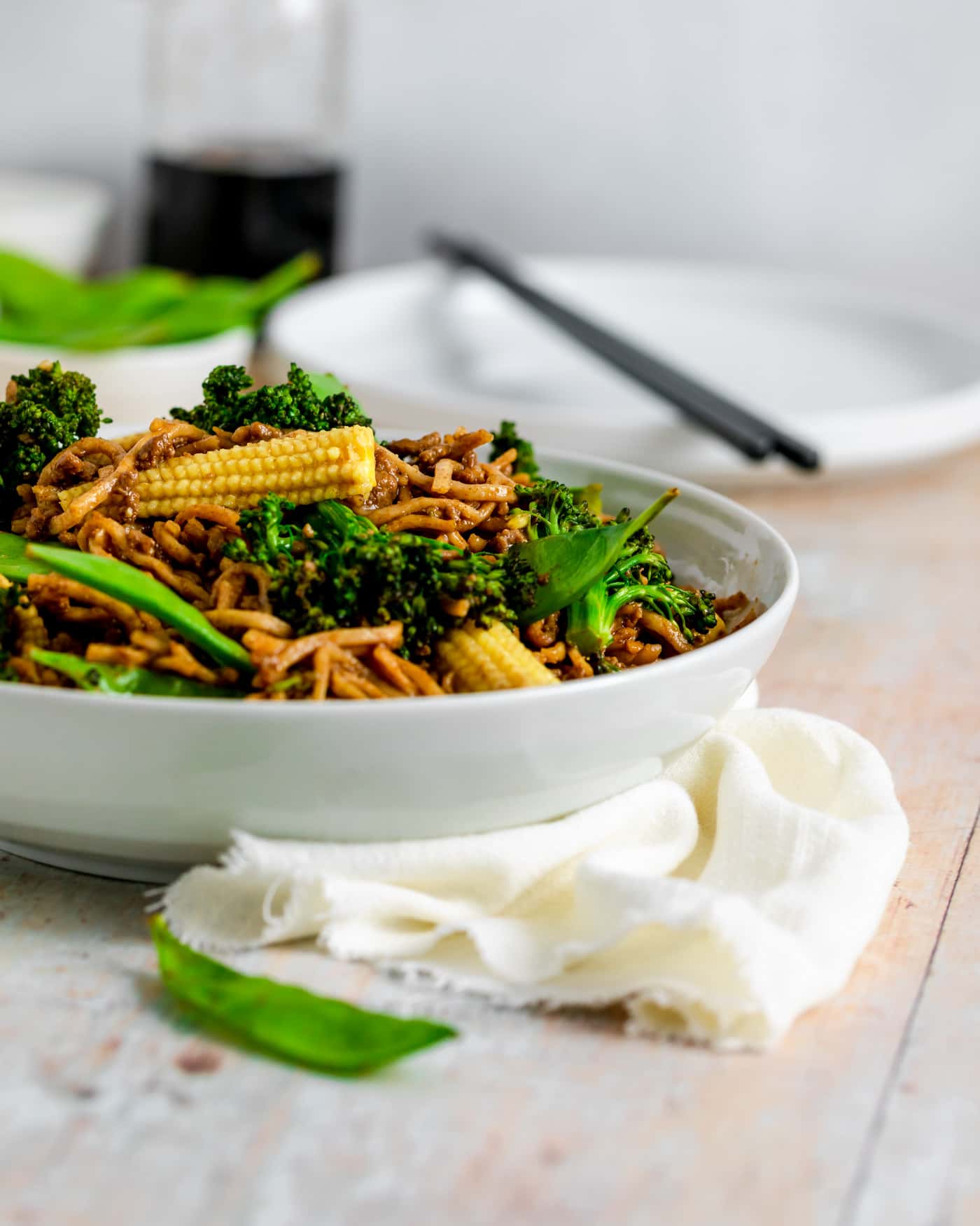 chinese style minced beef with noodles and green veg in a white serving bowl with a cream napkin swirled around the bottom of the dish.A white plate is in the background with chopsticks resting on it and a jar of soy sauce to the side and green mange tout in a small bowl