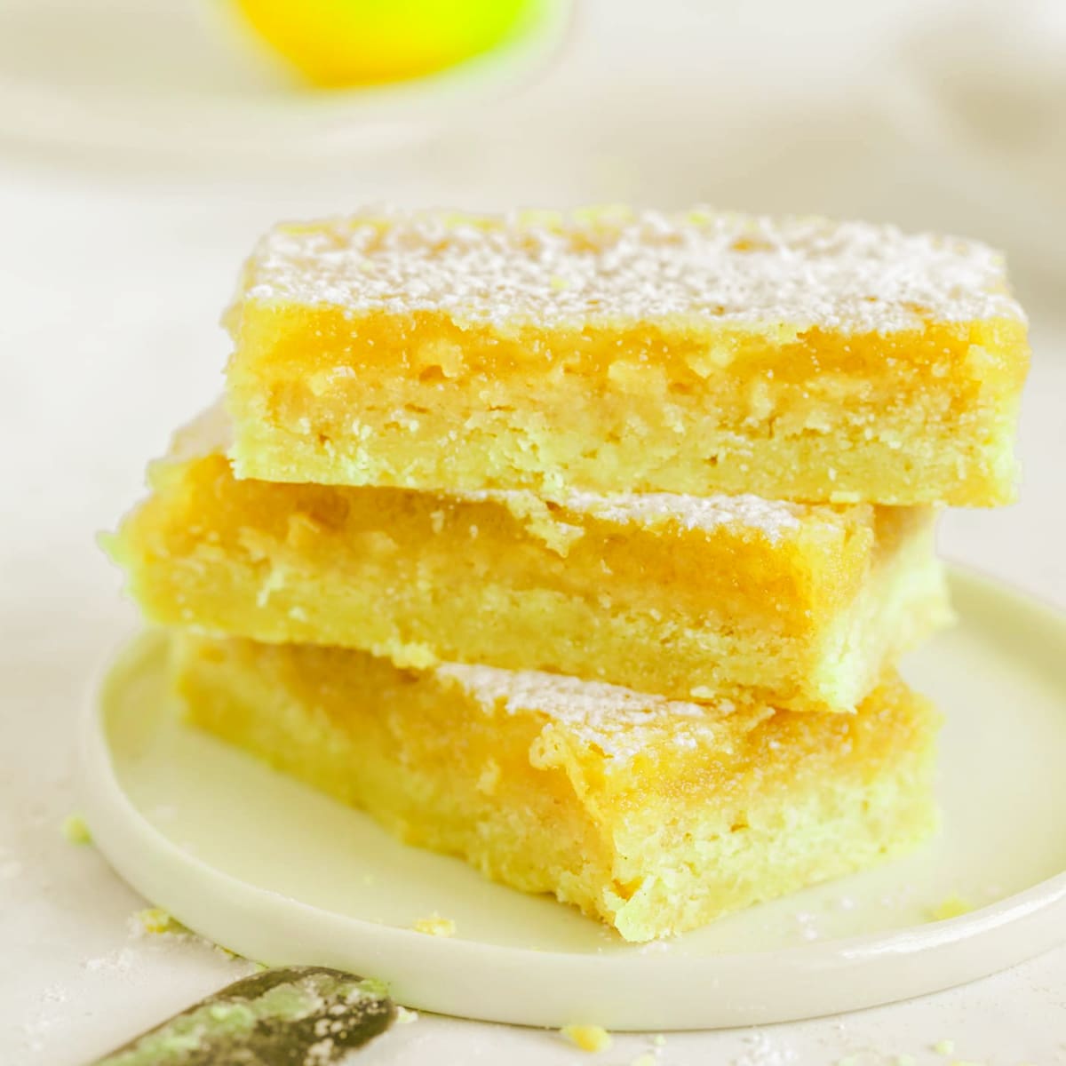 lemon bars stacked on top of each other on a small white lipped plate with crumbs and icing sugar scattered around a lemon is visible in the background