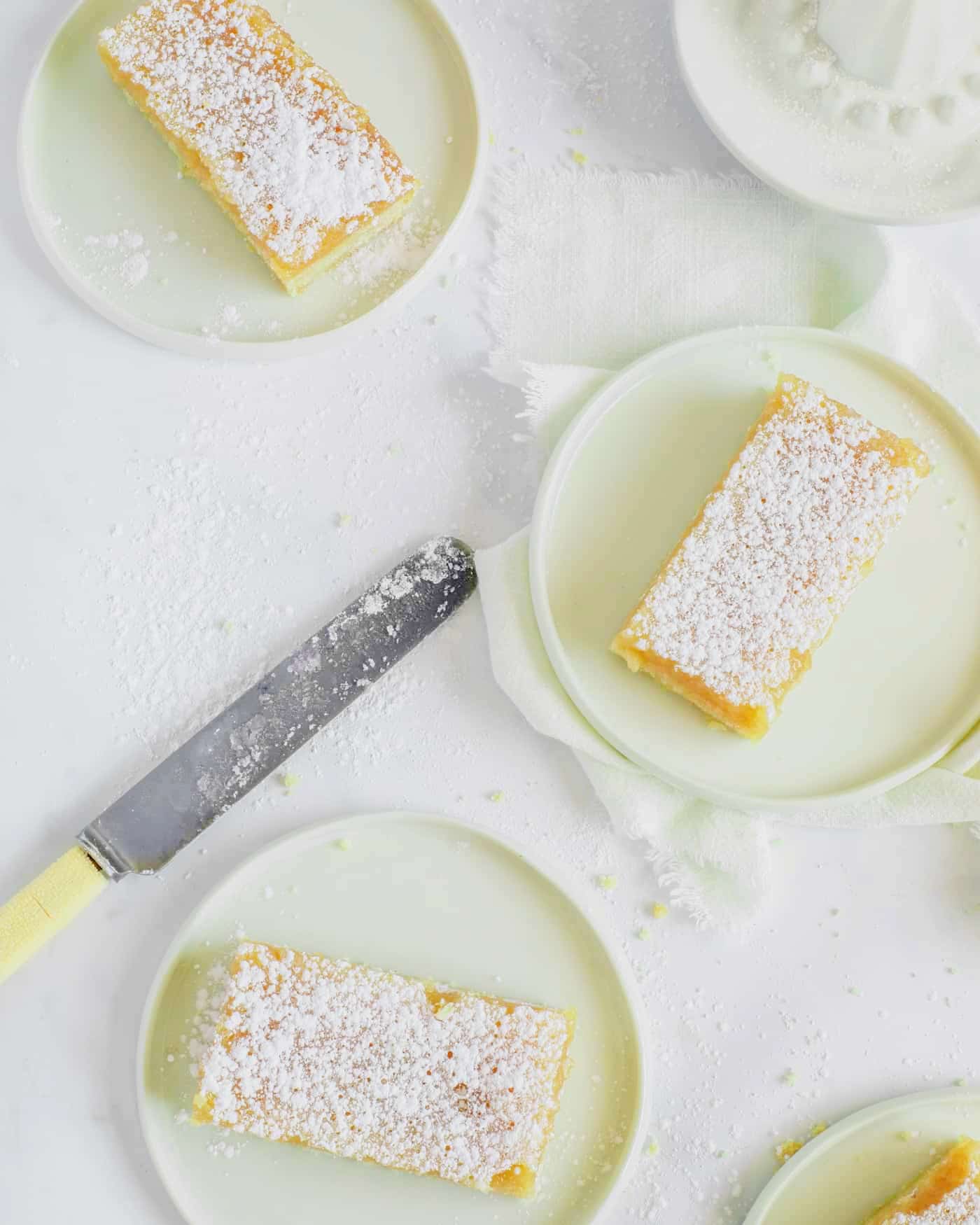 sliced lemon bars on small white lipped plates on a white work surface dusted with icing sugar. A yellow handled butter knife lies at an angle in the middle of the scene and a lemon juicer is at the top