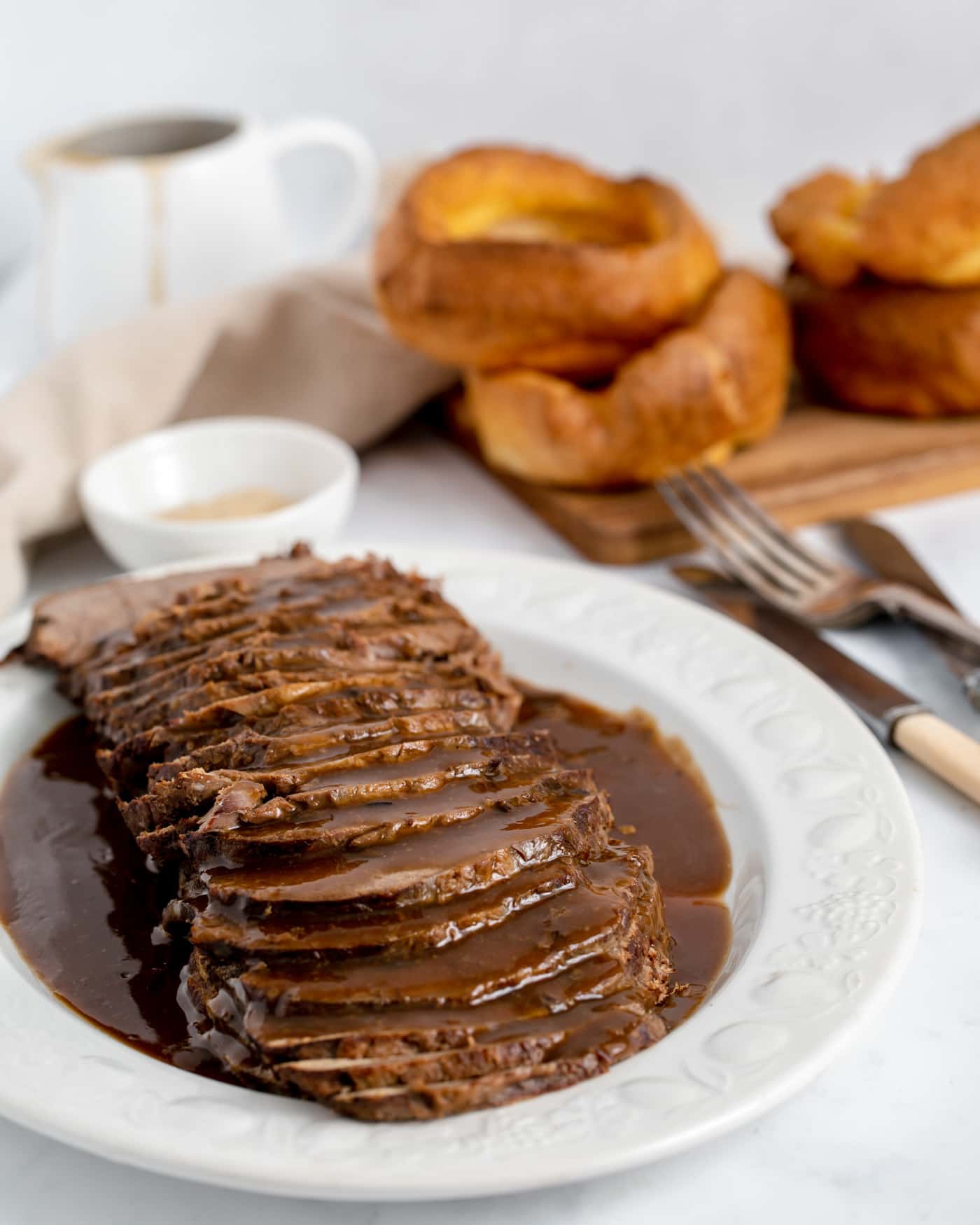 sliced roast beef with gravy on a large white serving platter with knives and forks to the side and yorkshire puddings at the back