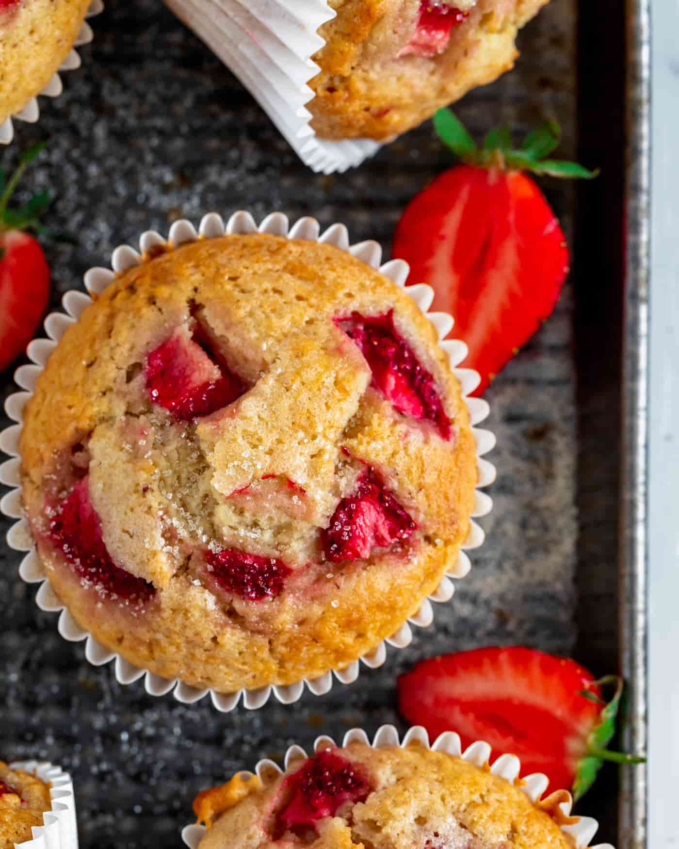 strawberry muffins on a metal baking tray with sliced strawberries scattered and two muffins lying on their sides