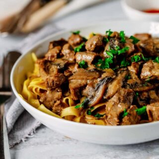 beef stroganoff on tagliatelle in a white bowl on a white work surface with a grey napkin to the side with a knife and fork on top. There is parsley sprinkled on top. A small white bowl of paprika is at the top of the bowl