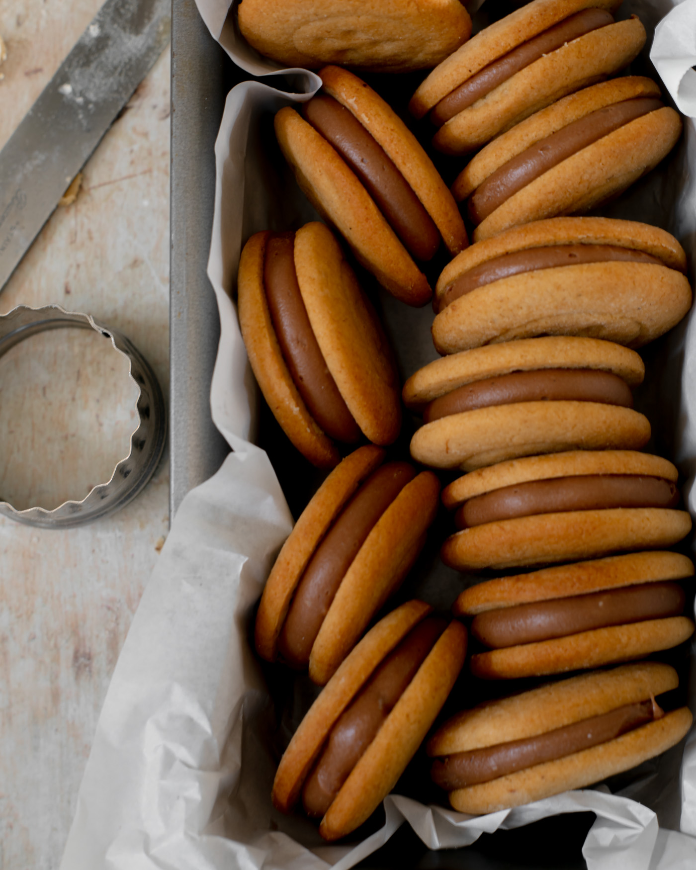 malted milk biscuits in a metal tin lined with white paper with a cutter to the side and a butter knife