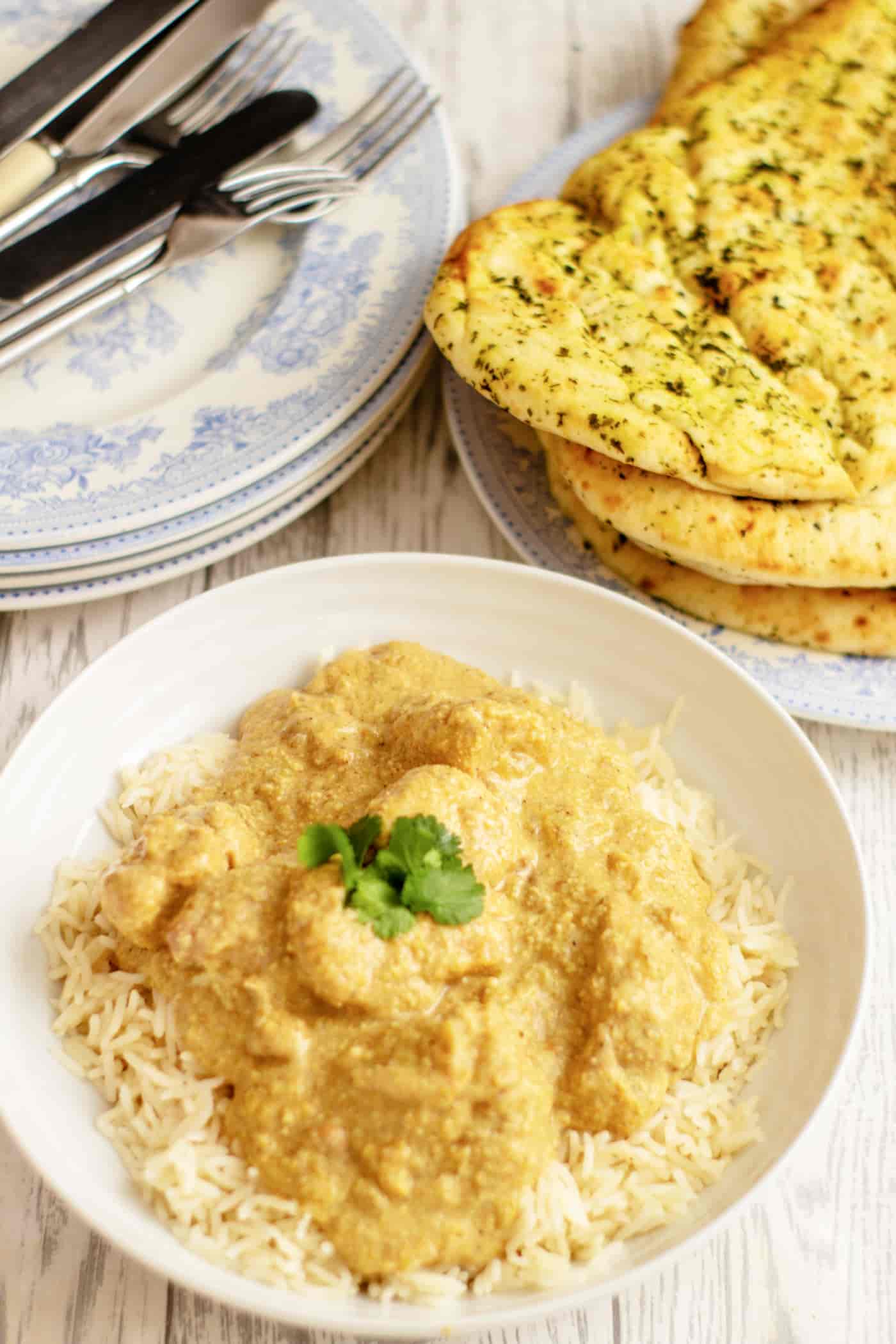 slow cooker chicken curry in a bowl with rice and a naan bread. Knives and forks rest on blue and white plates above