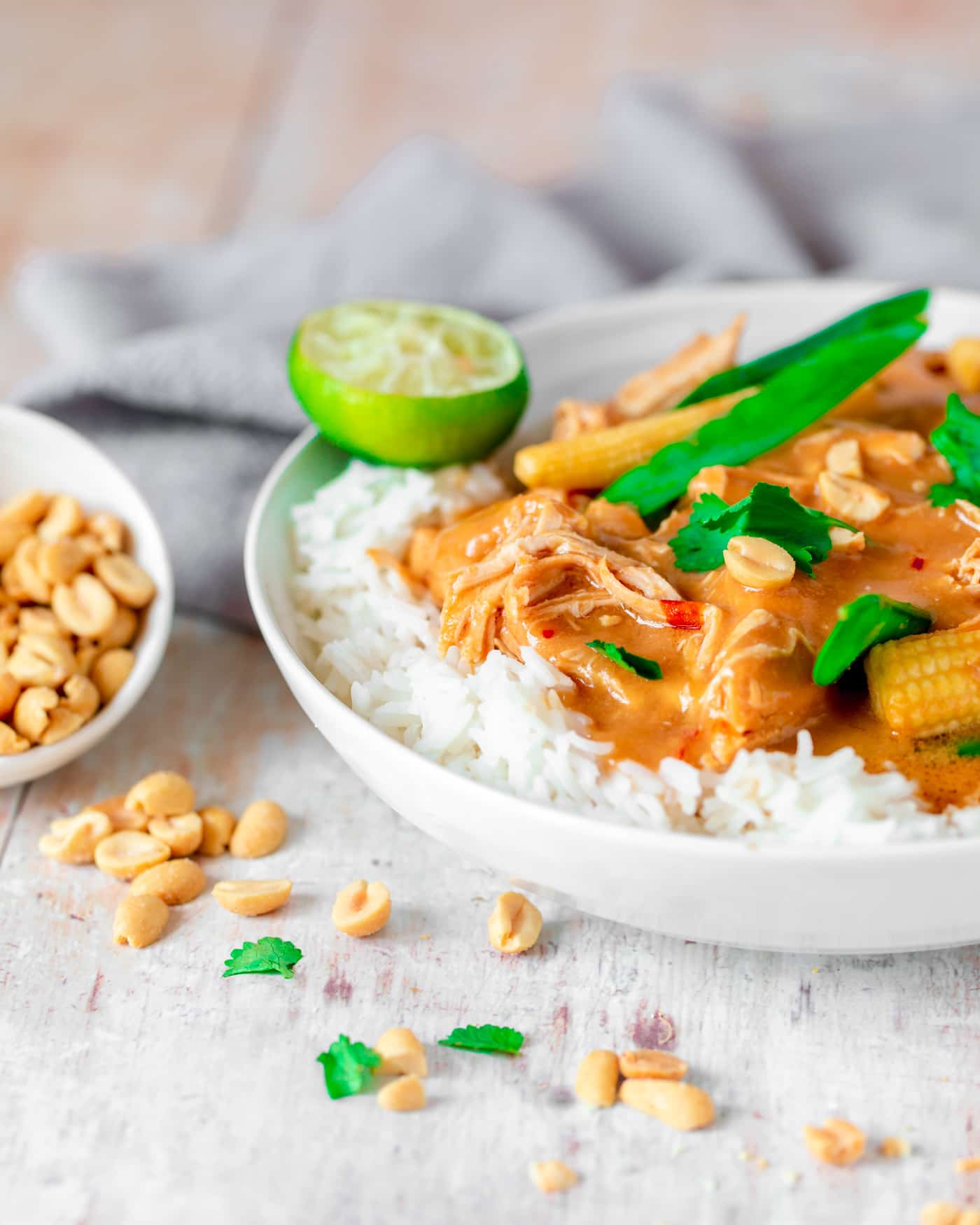 slow cooker peanut chicken in a white serving dish with white rice. Peanuts are falling out of a small white bowl and coriander is scattered at the front