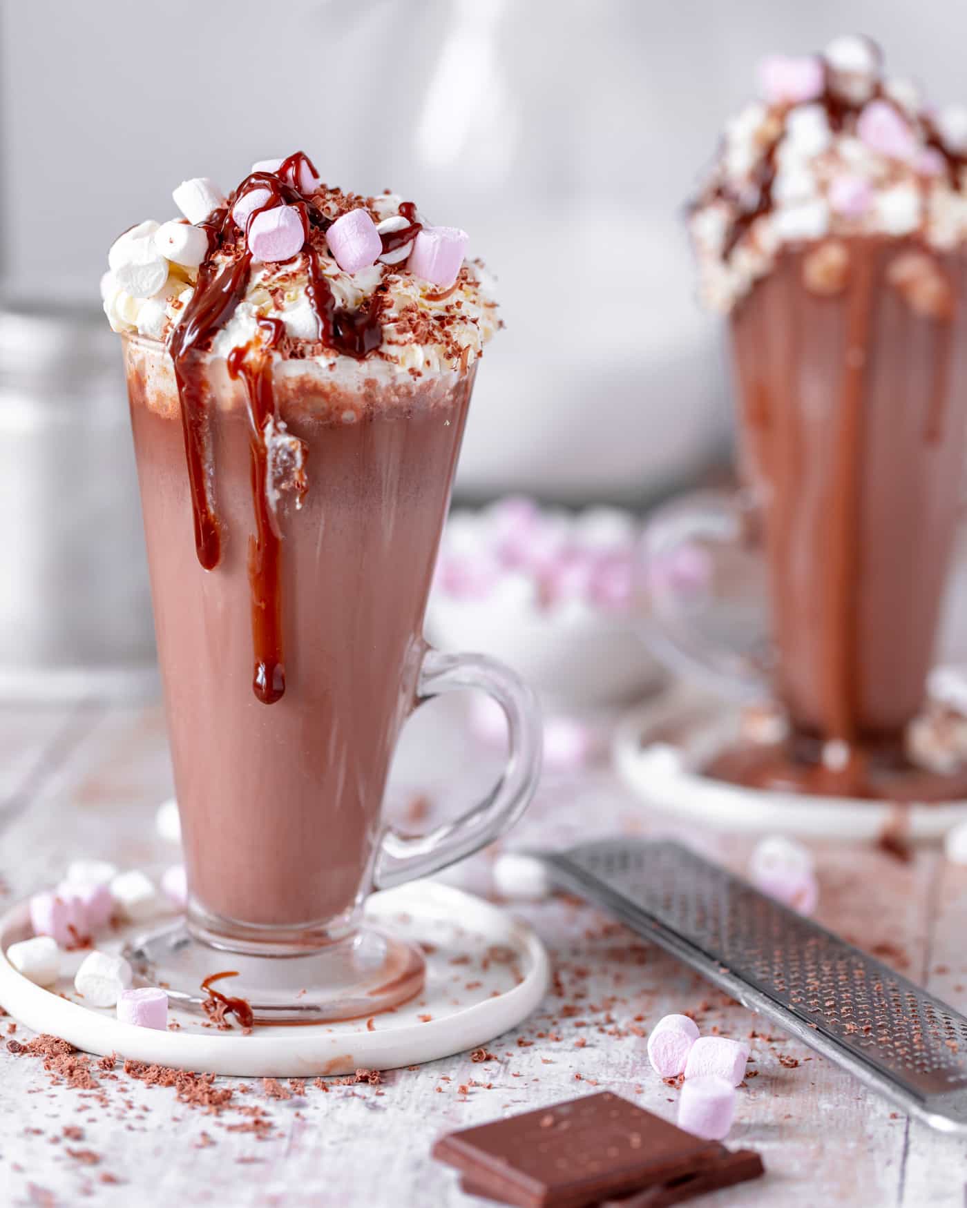 hot chocolate in a glass mug sat on a white small plate with mini marshmallows and grated chocolate chunks in the foreground. A long grater lies to the side and another glass tall mug of hot chocolate is in the background