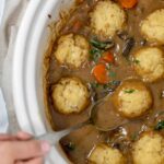 slow cooker beef stew with dumplings in a cream slow cooker pot with a hand holding a serving spoon out of the gravy with a dumpling balanced on it