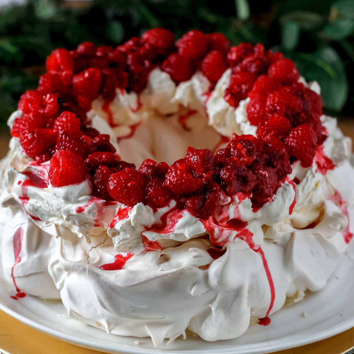 wreath of meringue piled high with whipped cream and red berries own a white serving plate and with dark greenery in the background on a table