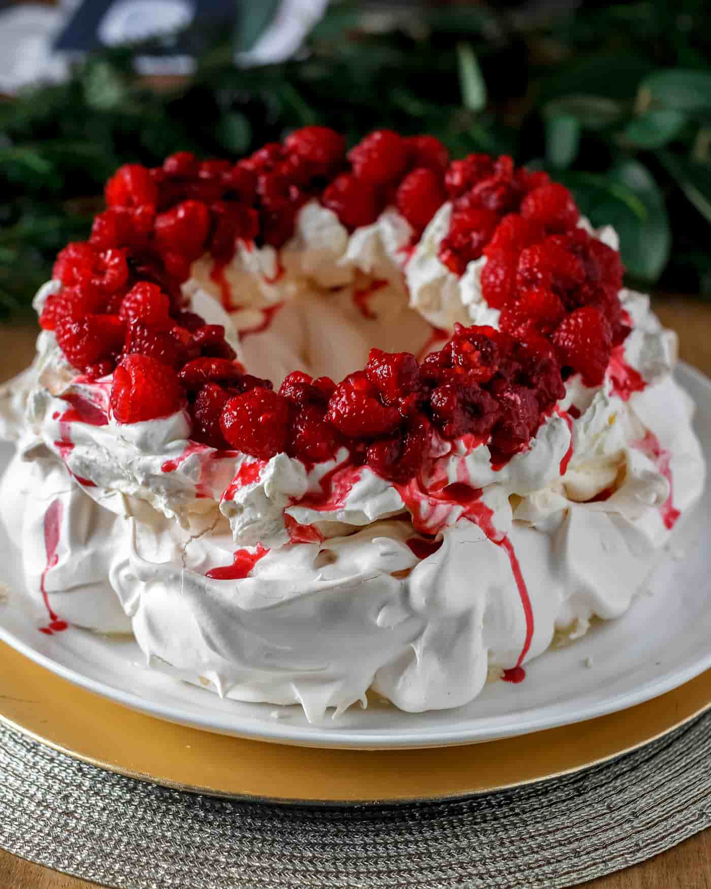 pavlova wreath topped with whipped cream and raspberries on a round white serving dish with greenery in the back ground