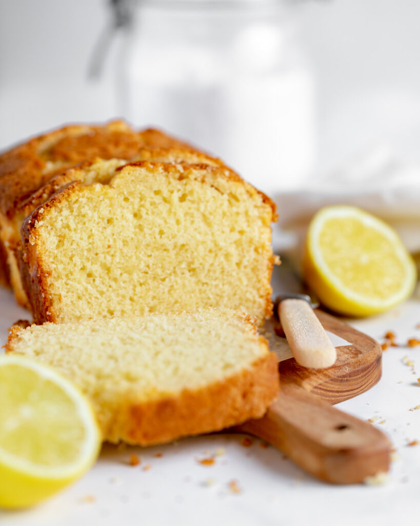 lemon loaf cake sliced on a wooden chopping board with a knife lying to the fore and a lemon sliced in half lying to the side and another to the front. Crumbs are scattered all around