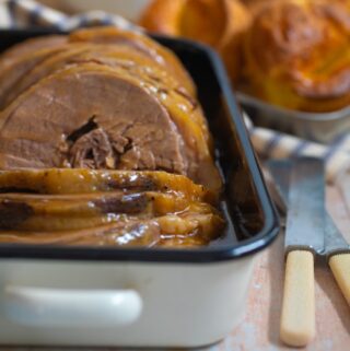 slow cooker brisket with gravy in a cream enamel dish inlaid with black. Knives and forks lie to the side. Yorkshire puddings are piled in the background and a grey striped napkin wraps around the back of the serving dish