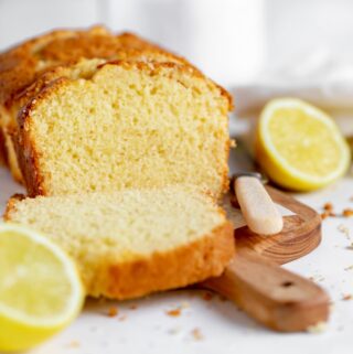 lemon loaf cake sliced on a wooden chopping board with a knife lying to the fore and a lemon sliced in half lying to the side and another to the fore. Crumbs are scattered all around on the white work surface