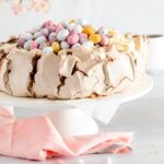 chocolate mini egg pavlova on a white cake stand with a pink napkin around the base and with pink flowers showing in the top left hand side of the picture