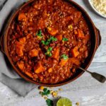 slow cooker red thai curry vegetarian raw ingredients in a brown round serving dish sitting on a grey linen napkin with sliced halves of lime on each side with a plate of rice