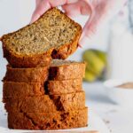 slices of banana loaf cake piled on top of each other with a hand lifting a piece off. Bananas are in the background and a small white bowl of ground cinnamon