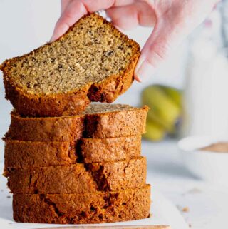 slices of banana loaf cake piled on top of each other with a hand lifting a piece off. Bananas are in the background and a small white bowl of ground cinnamon
