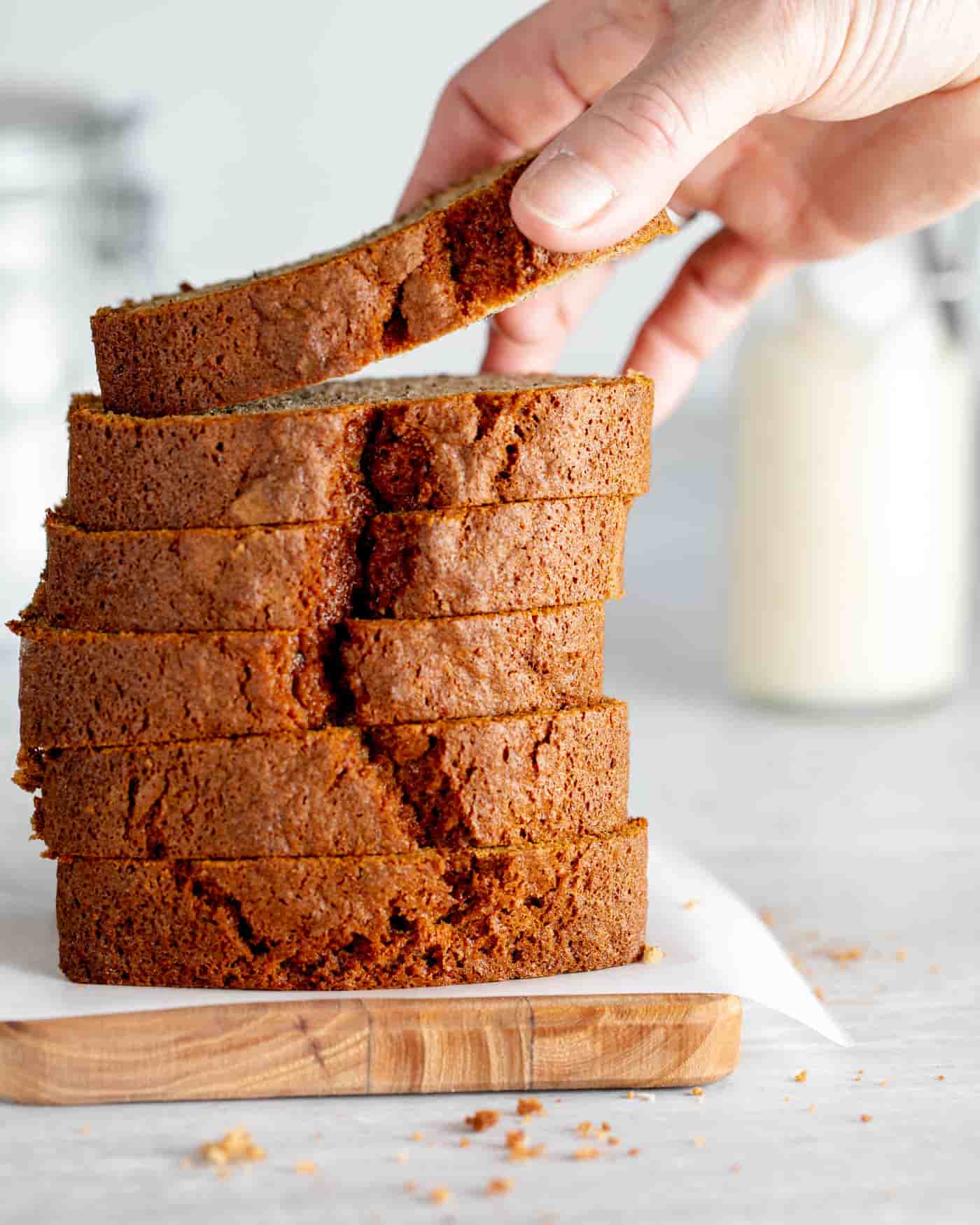 slices of banana bread loaf cake haphazardly piled on top of one another with bananas behind and a small white bowl of ground cinnamon to the side. A hand is lifting the top piece up