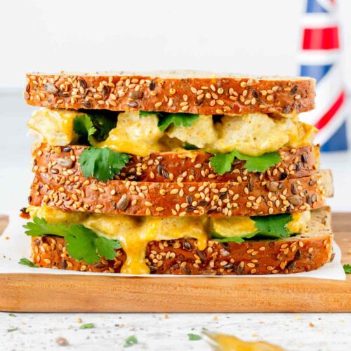 coronation chicken in sandwiches with coriander leaves poking out between the wholemeal seeded slices of bread all on a wooden board with tiny pieces of coriander and salt flakes scattered in the foreground along with a teaspoon covered in sauce. British flag is in the background