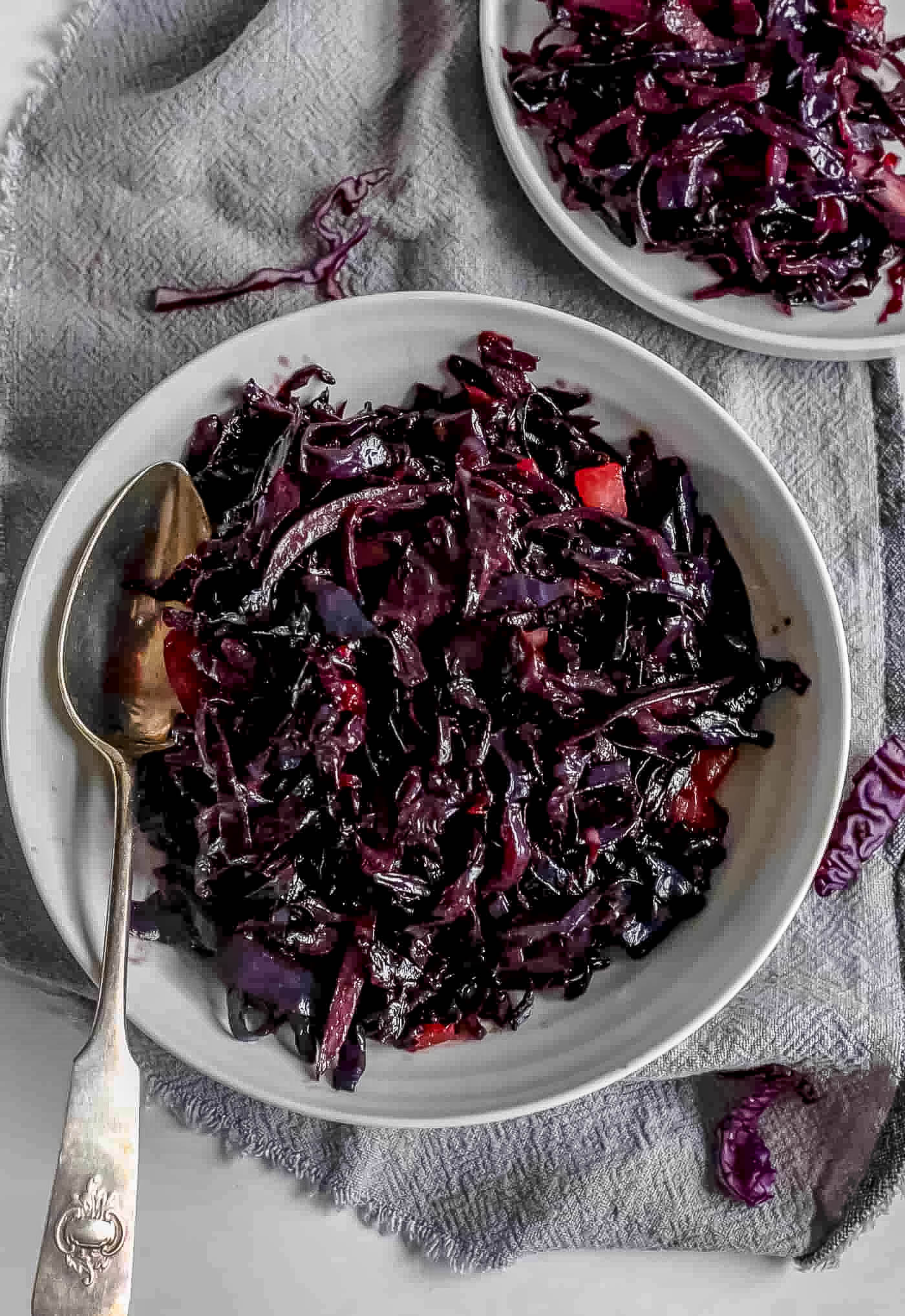 buttery braised red cabbage shredded in a white serving dish bowl with a large metal serving spoon resting on the side. On a grey napkin. A smaller white plate with red cabbage above