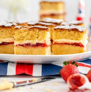 victoria sponge traybake on a white serving dish with british flag napkin below. A spoon ful of starwberry jam and a couple of strawberries are in the foreground with a knife laying to the fore on the other side