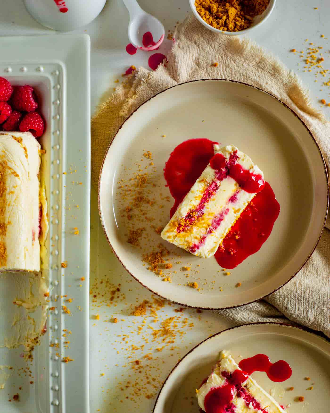 slices of cheesecake semifreddo on beige small lipped plates surrounded by ginger biscuit crumbs on a white surface . A long rectangular white plate with the rest of the semi freddo and some raspberries is to the side. A small white bowl is at the top half in sight