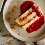 slice of cheesecake semifreddo on beige small lipped plate surrounded by ginger biscuit crumbs on a white surface and a beige napkin