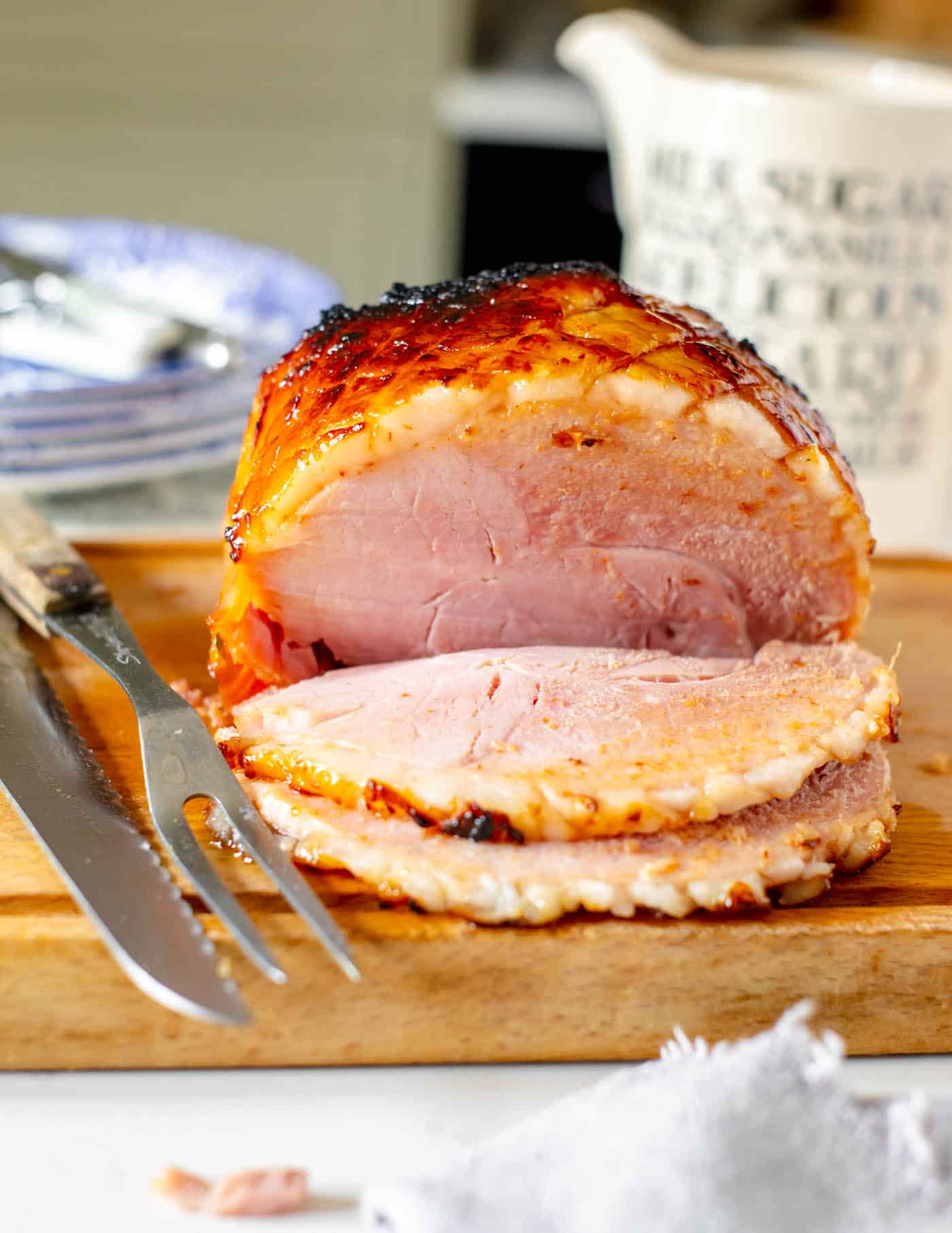 sliced gammon joint on a wooden chopping board with a carving knife and fork resting to the side. Plates are stacked behind with knives and forks on top.