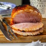 slow cooker gammon with brown sugar sliced on a wooden carving board with a carving knife and fork to the side. Blue and white plates lie behind next to a cream jug with black writing on it.