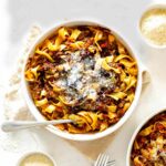 beef ragu with pappardelle in a white serving dish sprinkled with parmesan cheese and a fork resting on the side of the bowl on a beige napkin. Another bowl of ragu and pasta is partially visible
