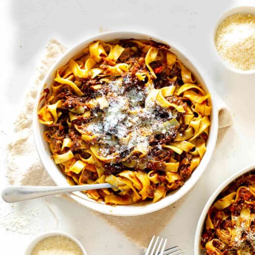 beef ragu with pappardelle in a white serving dish sprinkled with parmesan cheese and a fork resting on the side of the bowl on a beige napkin. Another bowl of ragu and pasta is partially visible