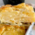 dauphinoise potatoes on a slice being held to the camera with a dish of dauphinoise below and a black slow cooker behind