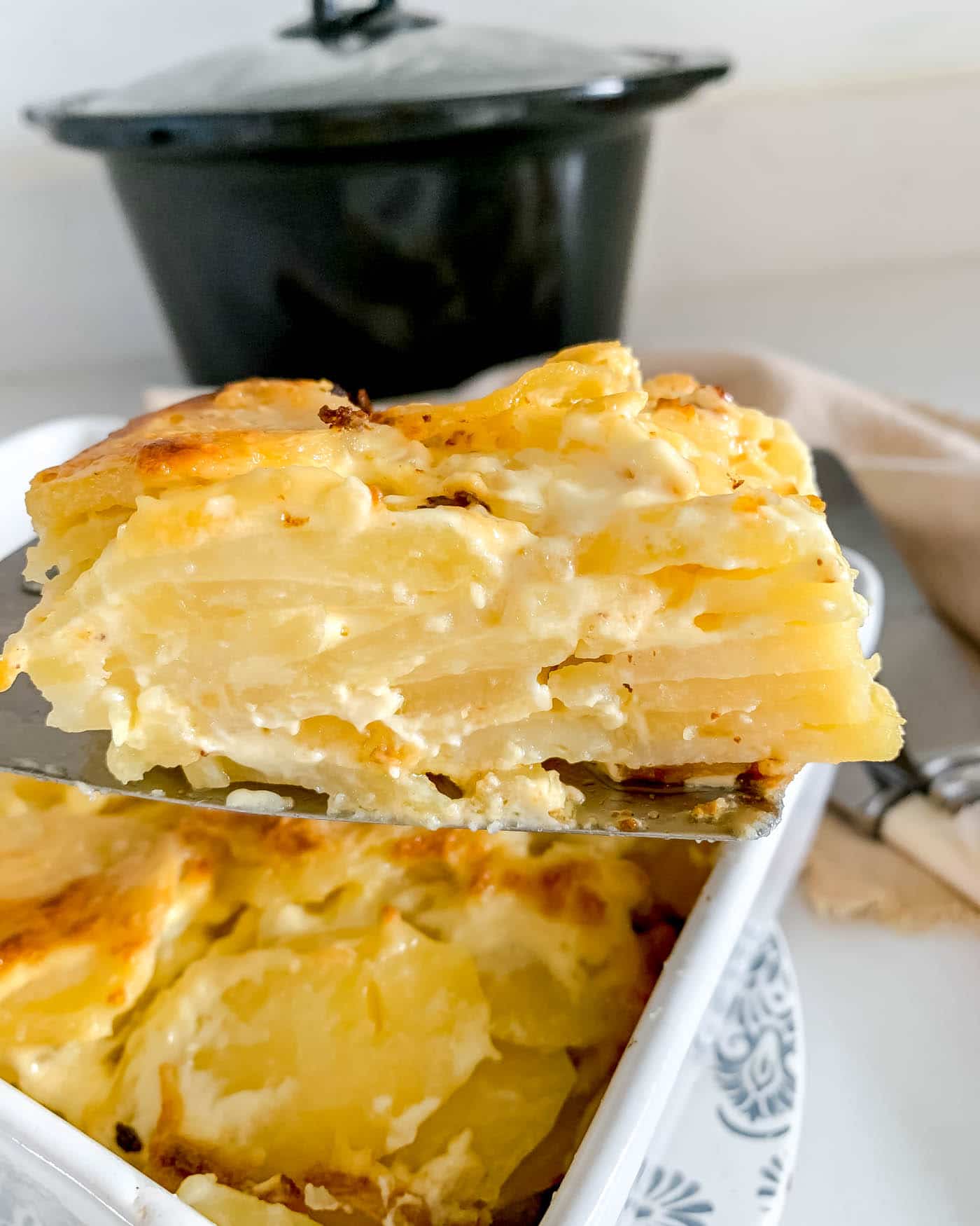dauphinoise potatoes on a metal slice held above a white dish of dauphinoise with a slow cooker in the background and knives and forks to the side