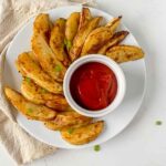 crispy potato wedges swirled round a white ramekin filled with ketchup on a white plate with a beige napkin curled around the plate