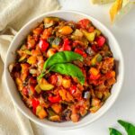 ratatouille served in a high lipped white round serving dish with two leaves of basil on top. Sliced baguettes to the side and a beige napkin curled around the dish