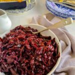 red cabbage in a white serving dish with a metal serving spoon resting on the side. Blue and white plates with knives and forks are visible to the back of the picture with a grey linen napkin curled around the dish