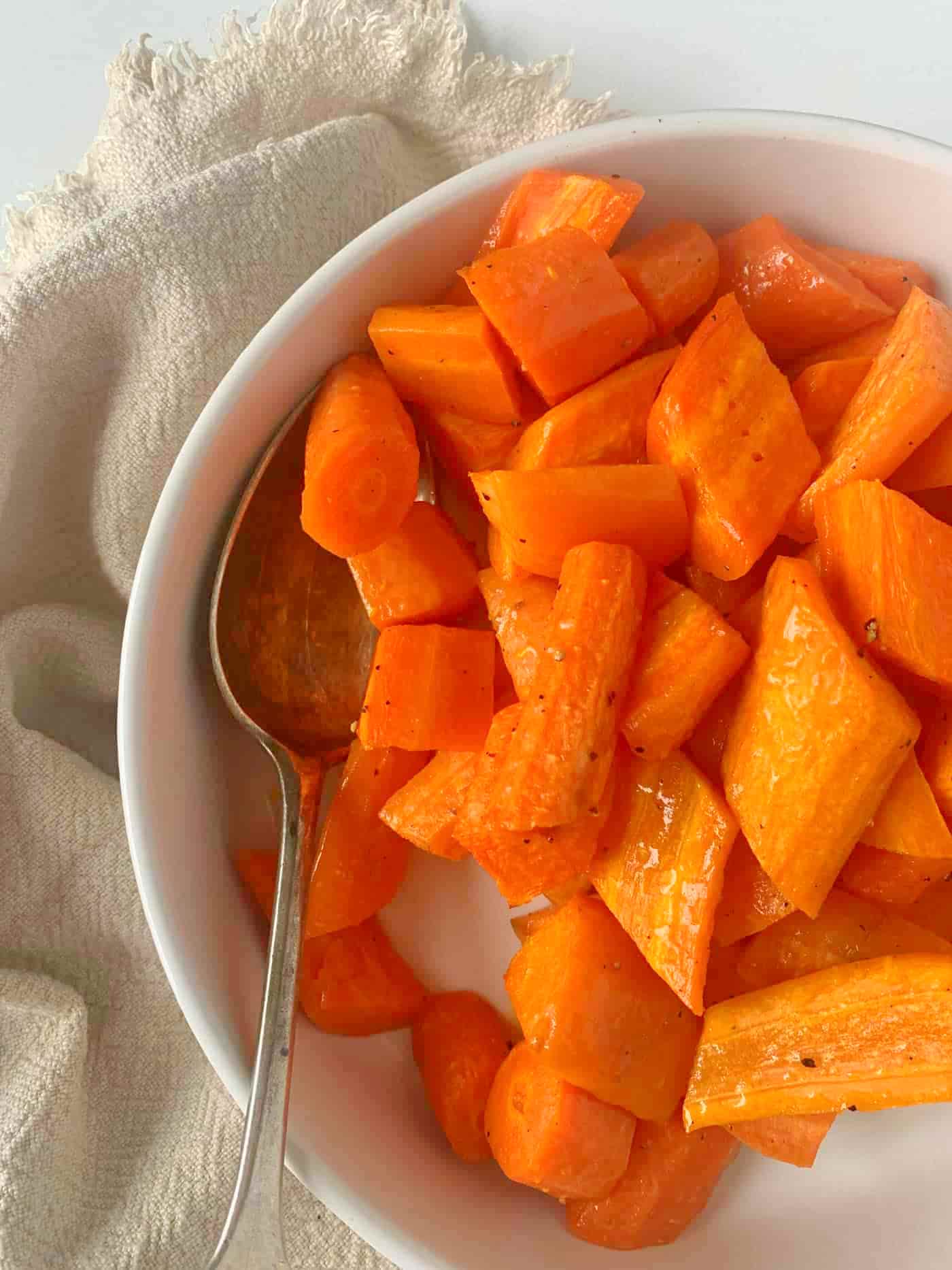 roasted carrots in a white serving dish with a serving spoon resting on the side with beige linen napkin around the dish