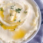 cheesy mashed potatoes in a white serving dish with melted butter drizzled over and herbs scattered over the top