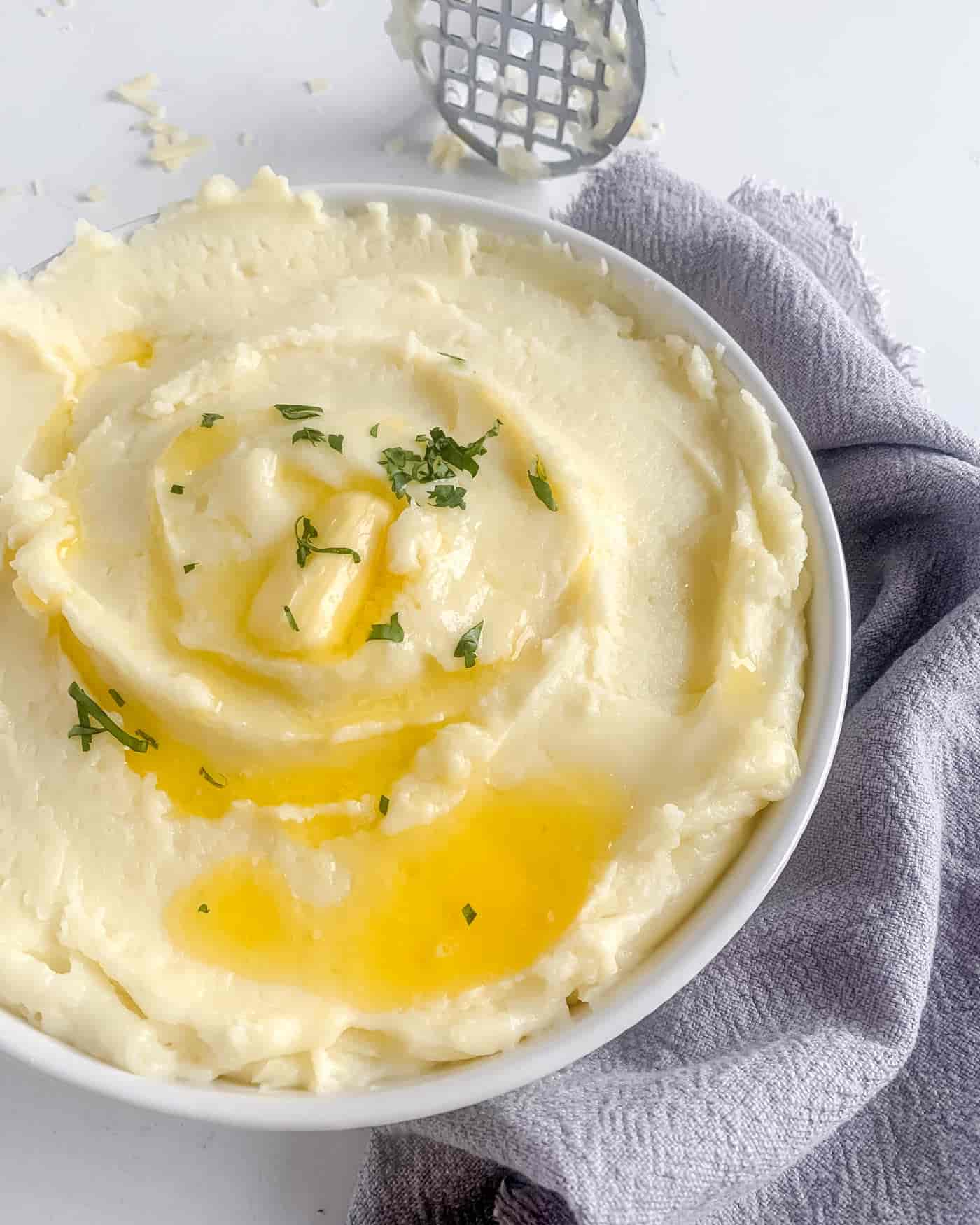 cheesy mashed potato in a white serving dish drizzled with butter and a few herbs. A grey napkin is wrapped around the dish and a potato masher is visible in the upper corner