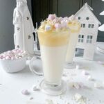 white hot chocolate in a glass mug topped with cream and mini marshmallows and grated white chocolate. White christmas decorations lie behind. Mini marshmallows and more grated white chocolate are scattered around the mugs