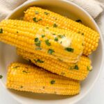 corn on the cob piled up in a white serving dish brushed with butter and parsley