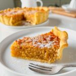 treacle tart slice on a white serving plate with a dessert fork resting on the side and the rest of the deep dish treacle tart behind
