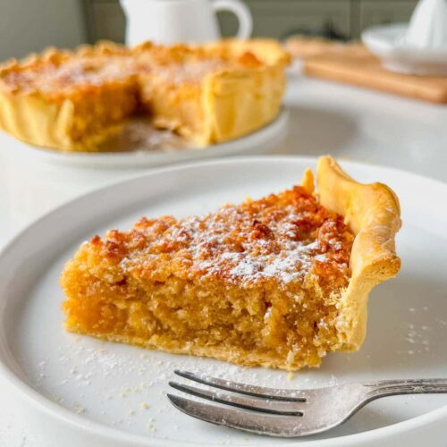 treacle tart slice on a white serving plate with a dessert fork resting on the side and the rest of the deep dish treacle tart behind