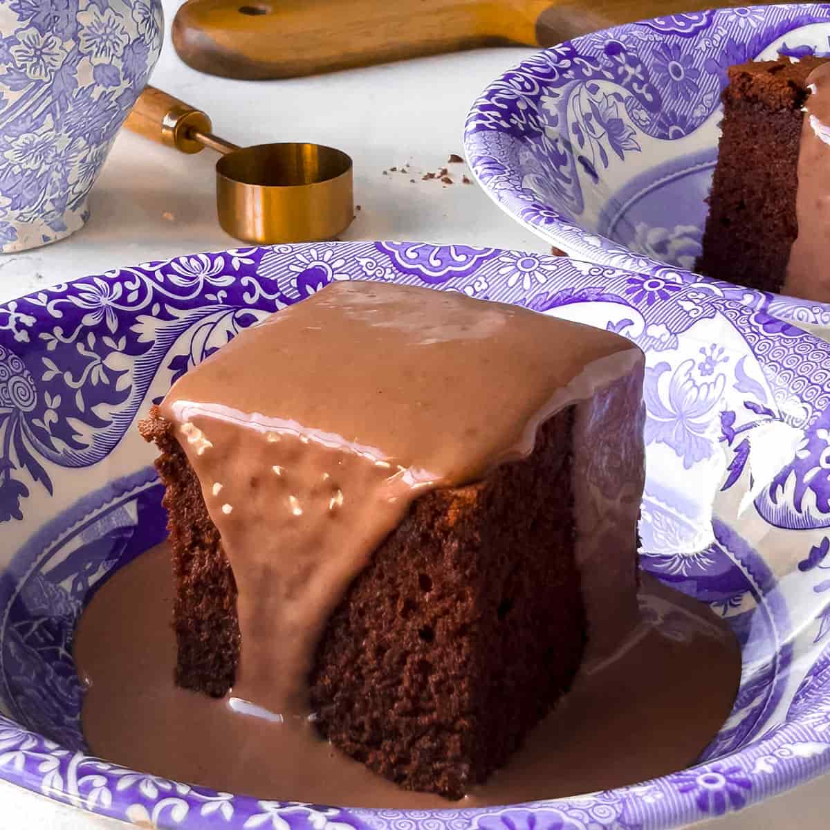 baked chocolate sponge pudding with chocolate custard poured over the sponge in a blue and white bowl with another bowl sitting behind it also with chocolate sponge and custard in