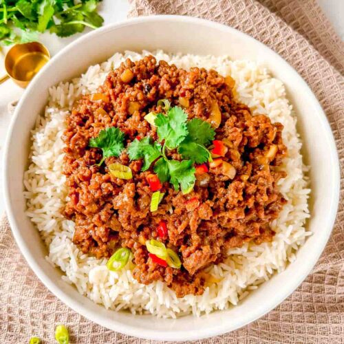 slow cooker asian minced beef on rice with garnish of coriander and sliced spring onions with coriander in the background and a copper measure