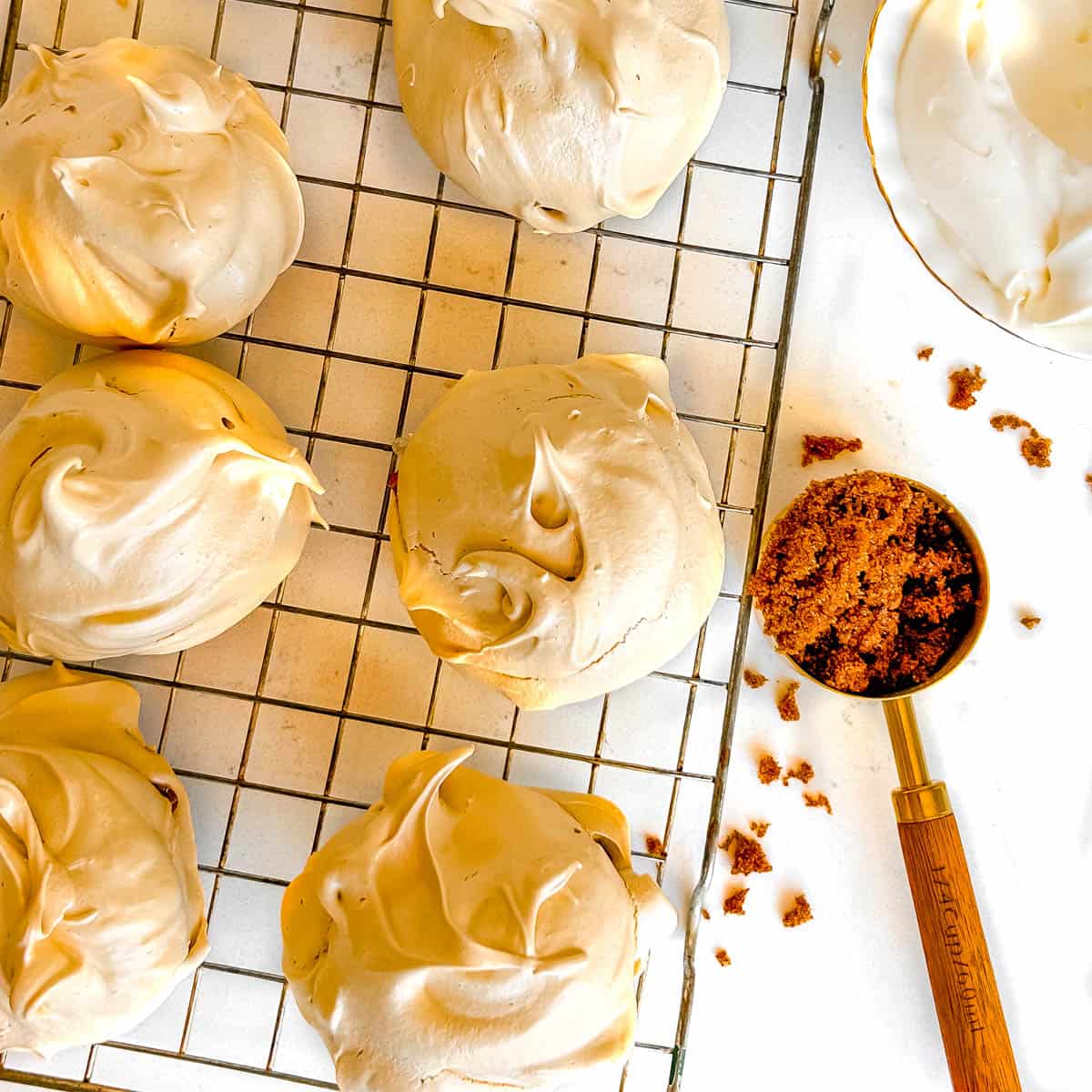 brown sugar meringues on a wire rack with a gold tbsp full of dark brown sugar and a bowl of whipped cream on the side in top corner