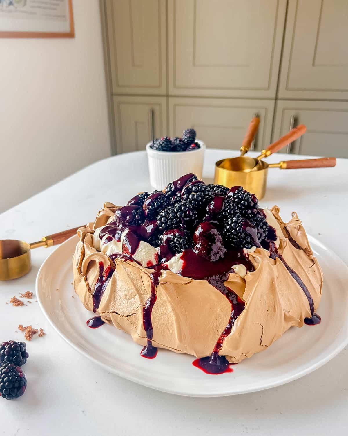brown sugar pavlova with cream and blackberries on a white serving plate with blackberries to the fore and brass measuring cups behind